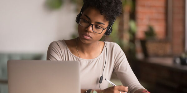 Focused mixed race woman wearing headphones watching webinar write notes study online with skype teacher, african female student learning language computer course on laptop listen translate lecture